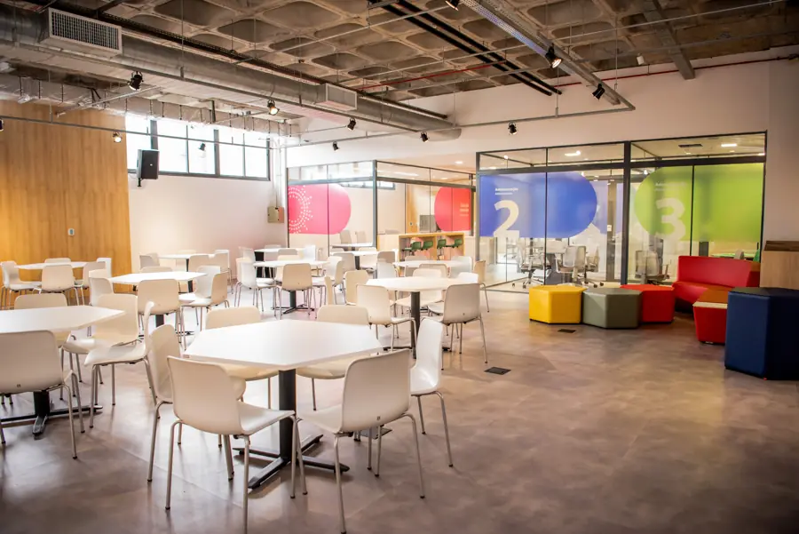 Espaço de convivência com mesas, pufes coloridos e sala de reunião envidraçada
